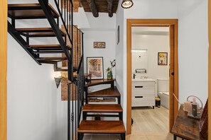Una escalera con escalones de madera y barandillas negras, que conduce a un cuarto con un lavamanos blanco y una mesa de madera.