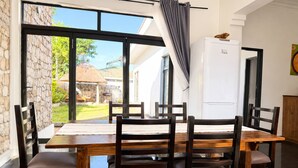A dining area with a large table and chairs, a white refrigerator, and a view of a garden through large windows. - Modern Vic Falls Home (Victoria Falls)