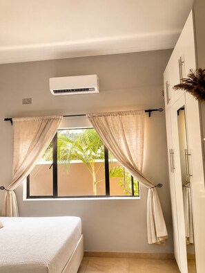 A bedroom with a bed, a window with curtains, an air conditioning unit, and a wardrobe. - Modern Vic Falls Home (Victoria Falls)
