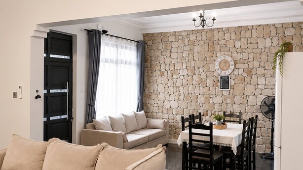 A modern living room with a beige sofa, a dining area with a table and chairs, and a stone wall. - Modern Vic Falls Home (Victoria Falls)