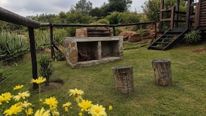 A stone-built outdoor fireplace with a brick surround, surrounded by a grassy area with wooden stumps and a wooden fence. - RIVERMAN CABIN COUNTRY LODGE (Tonteldoos)