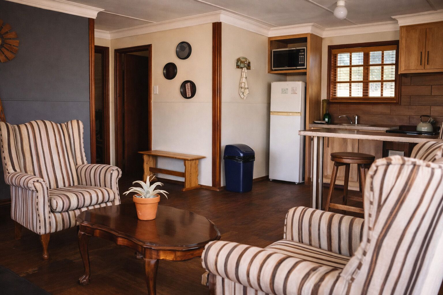 Uma sala de estar com poltronas listradas, uma mesa de centro, um vaso de planta, um banco de madeira pequeno e uma lixeira.