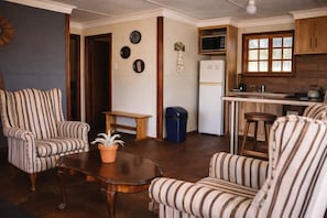 A living room with striped armchairs, a coffee table, a potted plant, a small wooden bench, and a trash bin. - RIVERMAN CABIN COUNTRY LODGE (Tonteldoos)