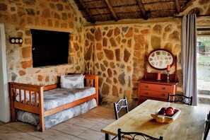 A rustic room with a stone wall, a wooden bed, a wooden table with a bowl of fruit, and a mirror.