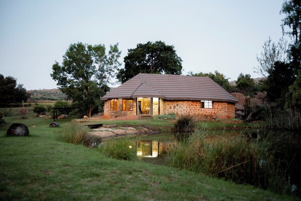 A stone house with a tiled roof, situated by a pond, surrounded by grass and trees.