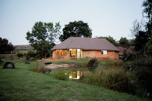 A stone house with a tiled roof, situated by a pond, surrounded by grass and trees. - RIVERMAN CABIN COUNTRY LODGE (Tonteldoos)