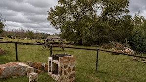 A fenced outdoor area with a stone structure, possibly a shelter, and a thatched-roof hut in the background. - RIVERMAN CABIN COUNTRY LODGE (Tonteldoos)