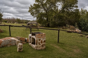 A fenced outdoor area with a stone structure, possibly a shelter, and a thatched-roof hut in the background. - RIVERMAN CABIN COUNTRY LODGE (Tonteldoos)