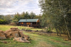 A rustic cabin with a green roof, surrounded by a grassy field and large rocks. - RIVERMAN CABIN COUNTRY LODGE (Tonteldoos)