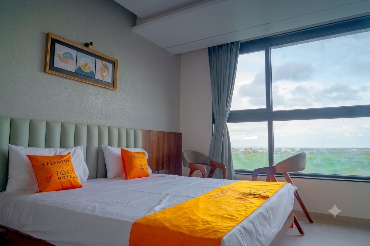 Habitación de hotel con cama, una silla, ventana con vistas y un cuadro en la pared.