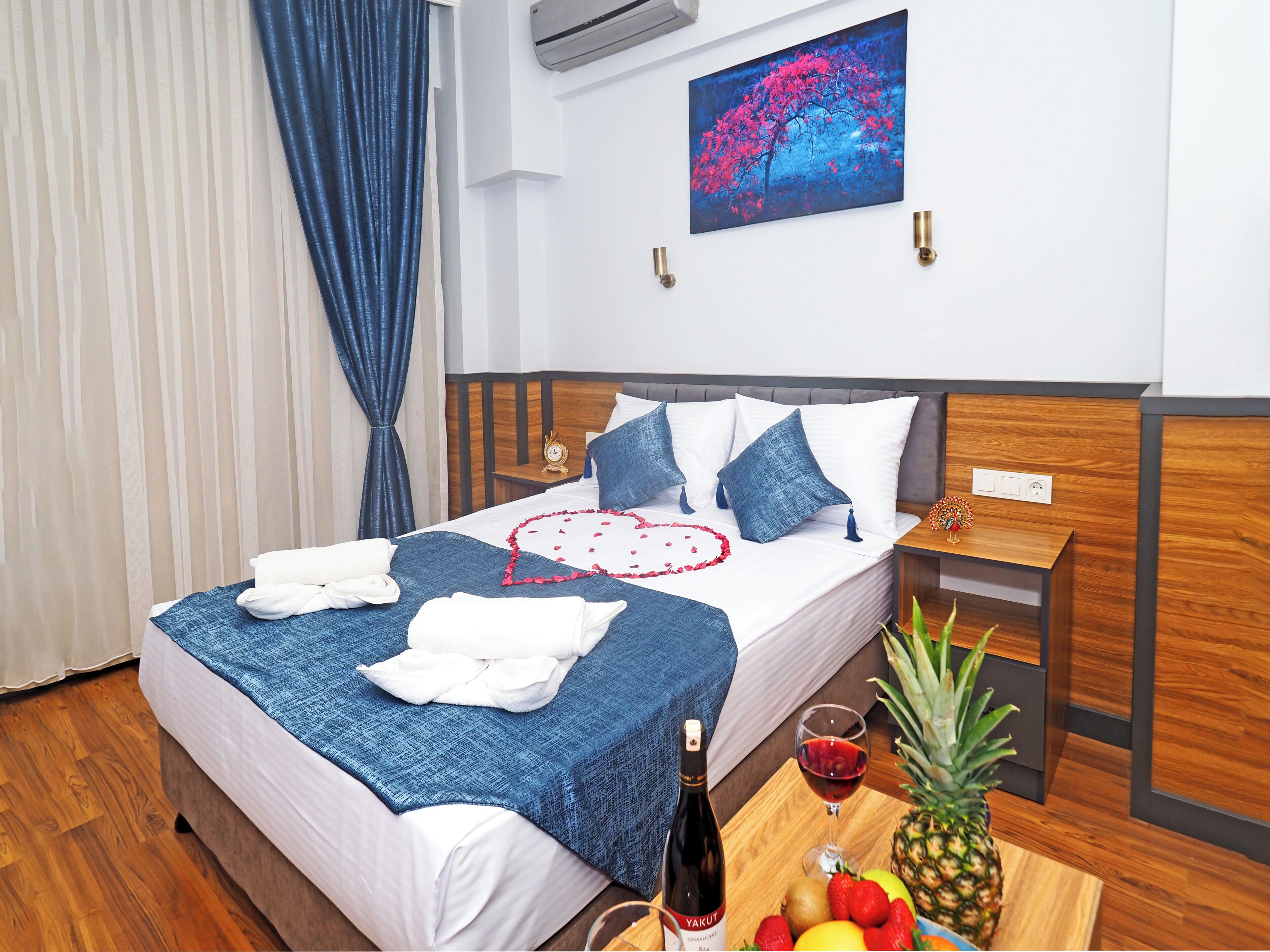 A neatly made bed with a blue blanket, white pillows, and a decorative heart on the bedspread. There is a wooden nightstand with a clock and a small plant. A bottle of wine and a fruit bowl are on the nightstand.
