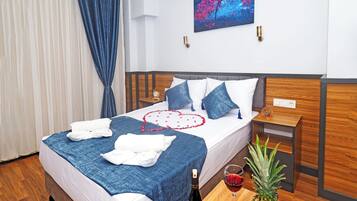 A neatly made bed with a blue blanket, white pillows, and a decorative heart on the bedspread. There is a wooden nightstand with a clock and a small plant. A bottle of wine and a fruit bowl are on the nightstand.