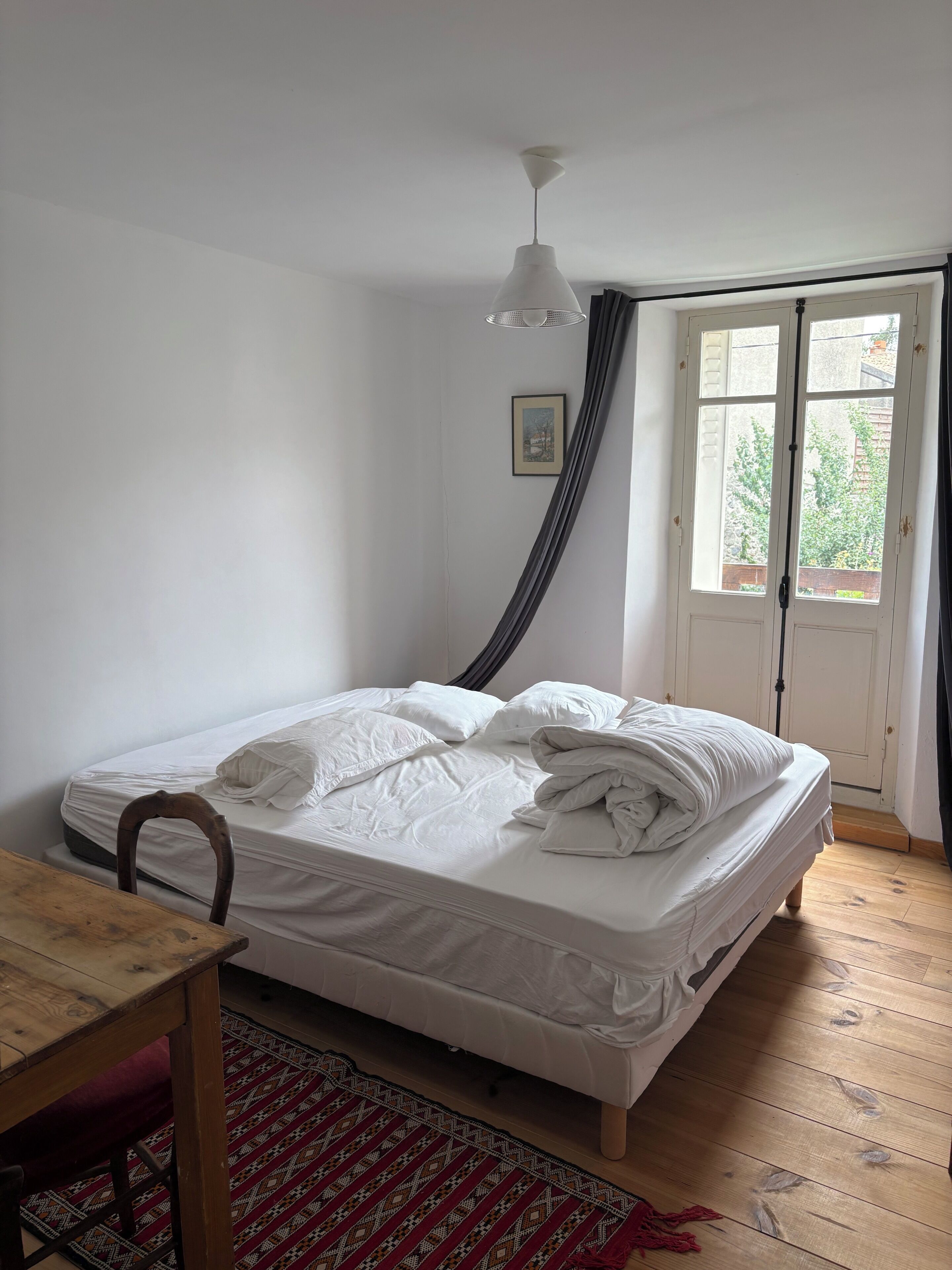Un lit simple avec du linge de lit blanc, une table en bois, un tapis à motifs et une fenêtre avec des rideaux.