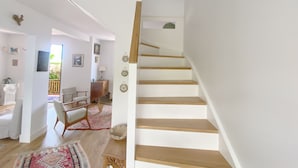 Eine Treppe mit Holzdielen und einer weißen Wand. Eine Tür führt in einen anderen Raum mit einem Stuhl und einem Tisch.
