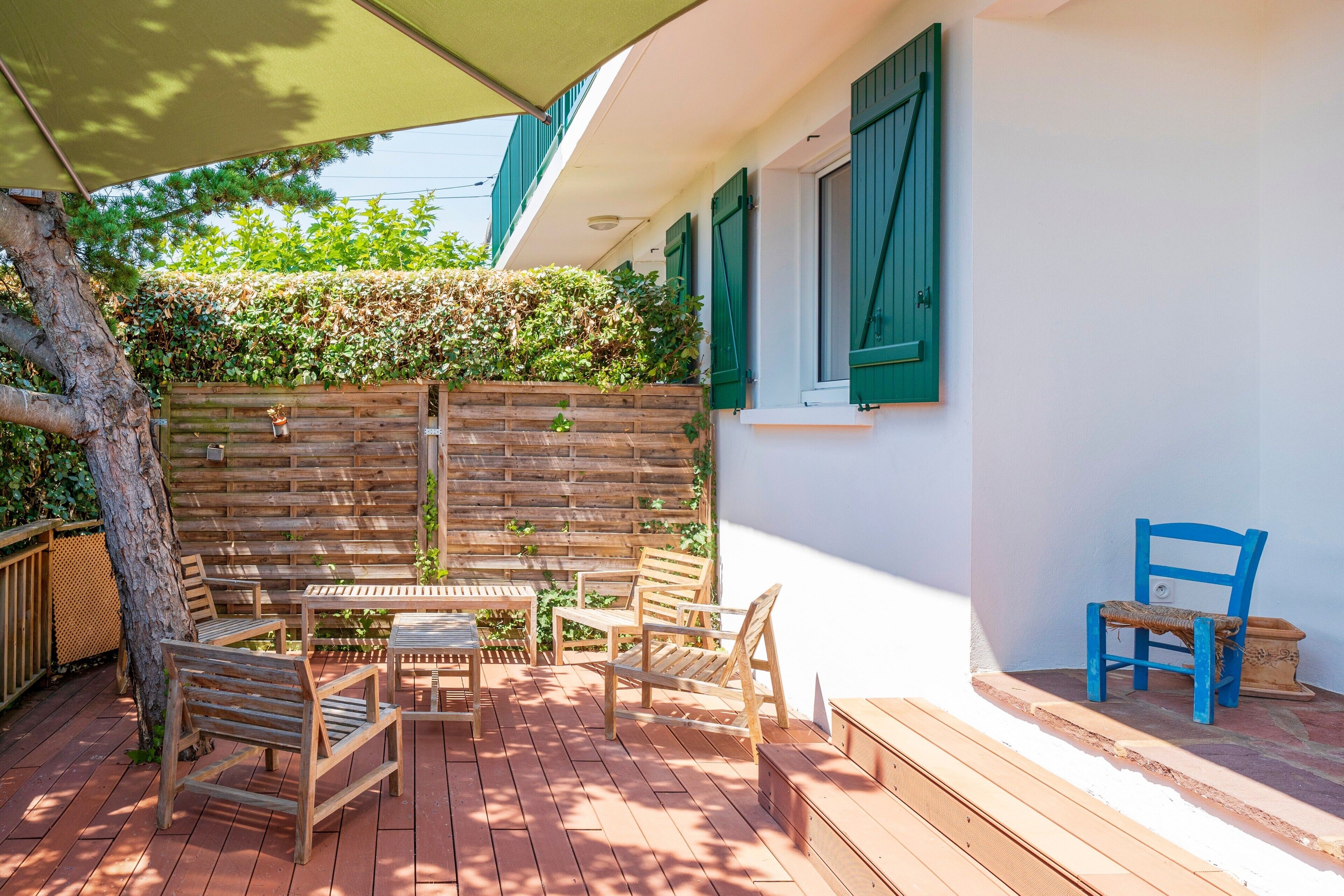 Una terraza de madera con mobiliario exterior, una valla de madera y un muro con contraventanas verdes.