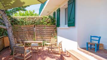 Una terraza de madera con mobiliario exterior, una valla de madera y un muro con contraventanas verdes.