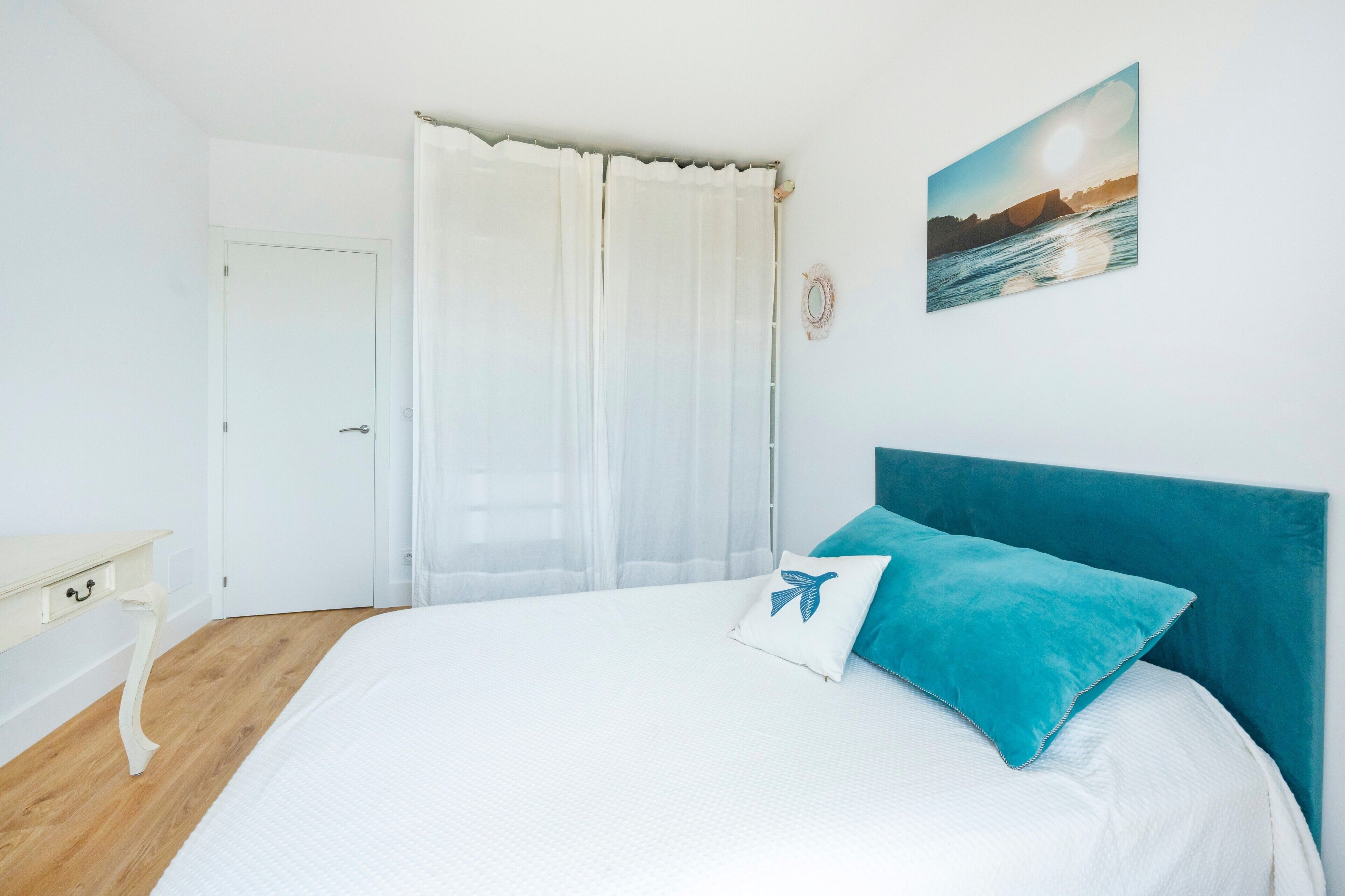 Ein Schlafzimmer mit einem Bett, einem weißen Nachttisch, einer Tür und einem Fenster mit durchsichtigen Vorhängen.