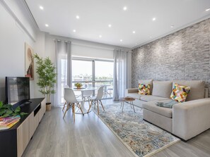 A modern living room with a sofa, a dining table with chairs, a TV, and a large window with curtains. - Solmonte Apartment - Pool Access, Beach Close (Portimão)