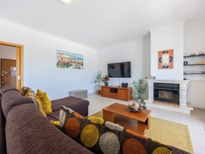 A modern living room with a sofa, coffee table, TV, fireplace, and a view of the outdoors. - Casa Iguana - Village Apartment with Shared Pool (Budens)