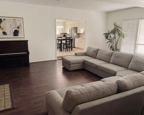 Ein Wohnzimmer mit einem Ecksofa, einem Klavier und einem Essbereich im Hintergrund.