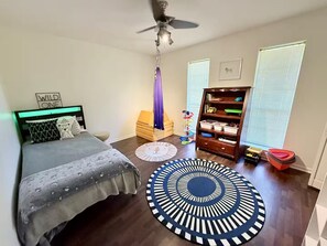 Ein Schlafzimmer mit einem Bett, einem Deckenventilator, einem Bücherregal und einem Fenster mit Jalousien.