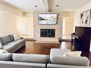 A living room with a fireplace, a sofa, a piano, and a television mounted on the wall. - Sensory-Friendly Family Retreat - Close To Park - 4-bedroom bungalow with AC (Texarkana)
