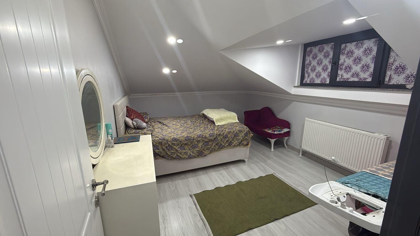 Un dormitorio con cama, una silla, un radiador y una mesita de noche con espejo.