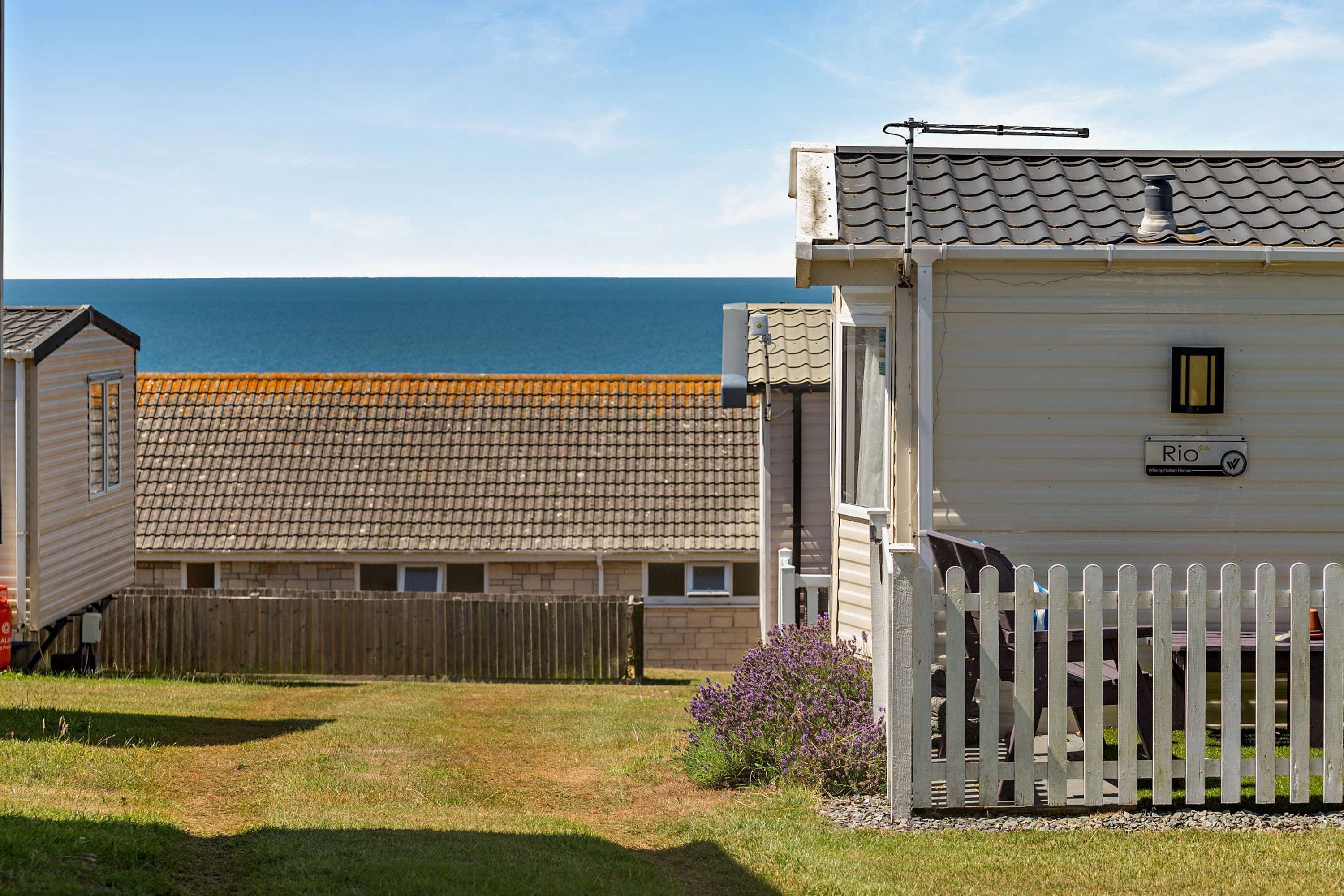 Une propriété située sur la côte, avec une clôture en bois peinte en blanc, un petit jardin agrémenté de fleurs violettes et une vue sur la mer.