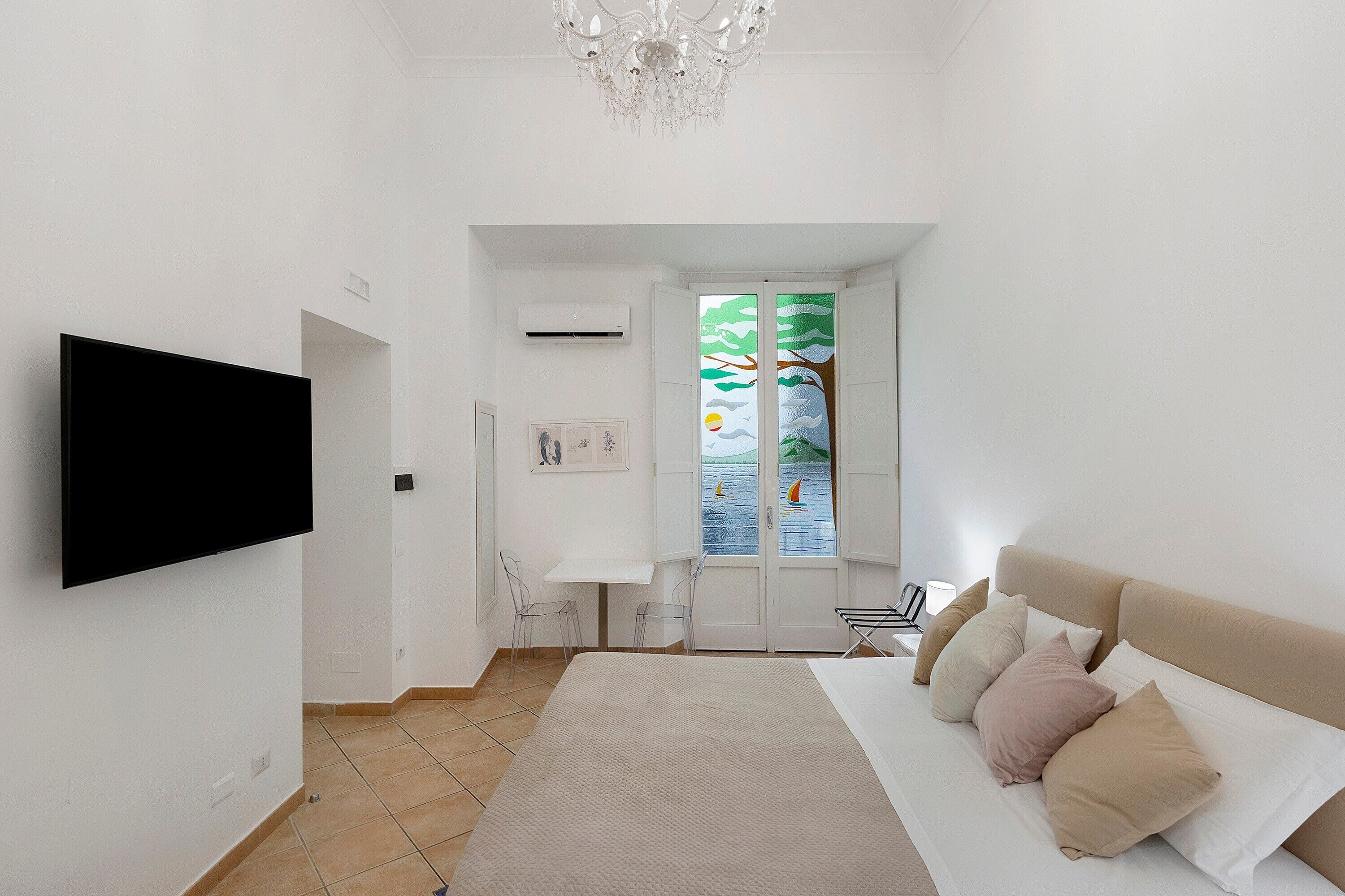 A bedroom with a bed, a TV, a chandelier, and a window with a view of the sea.