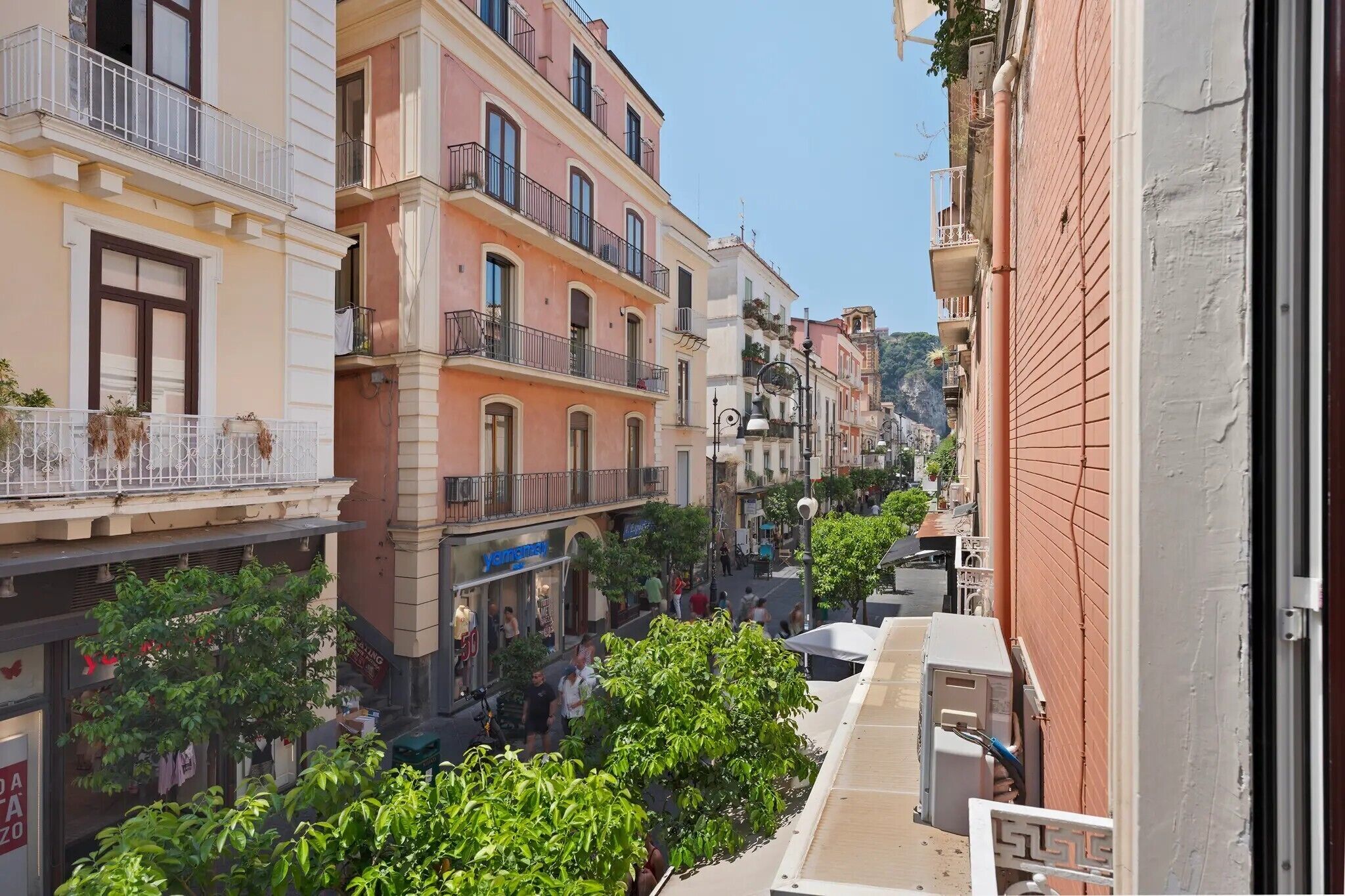 A narrow street with multi-story buildings, balconies, and a shop with a visible sign.