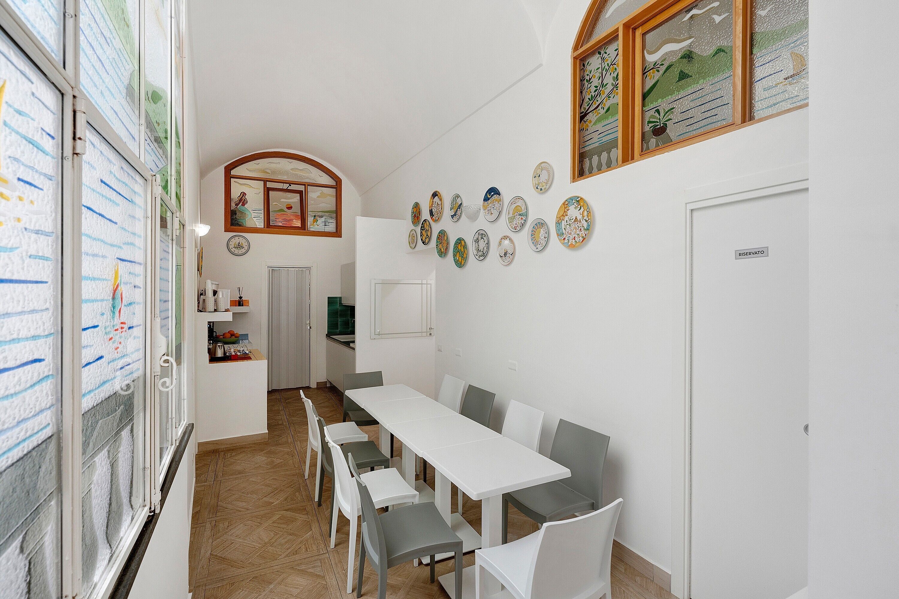 A dining area with a long table and chairs, a kitchen in the background, and decorative plates on the wall.