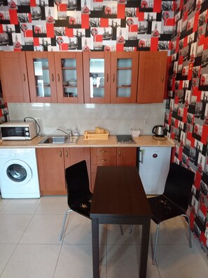 Eine kompakte Küche mit Holzausstattung, einer Mikrowelle, einem Spülbecken und einem kleinen Kühlschrank. Es steht ein Tisch mit zwei Stühlen und eine Waschmaschine in der Ecke.