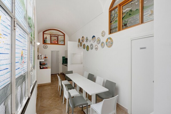 A dining area with a long table and chairs, a kitchen in the background, and decorative plates on the wall.