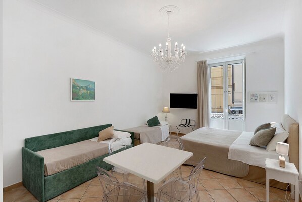 A modern hotel room with a green sofa, a white table, and a bed. There is a chandelier, a painting on the wall, and a window with curtains.