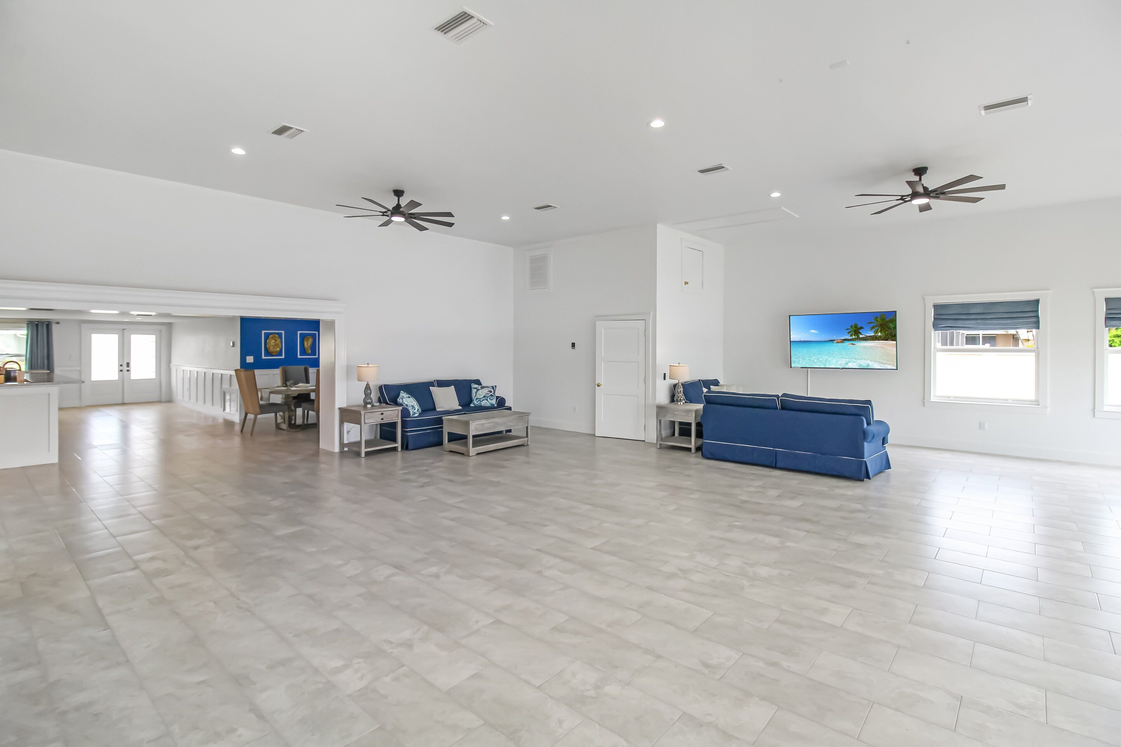 A spacious living area with a large TV, ceiling fans, and a kitchen area in the background.