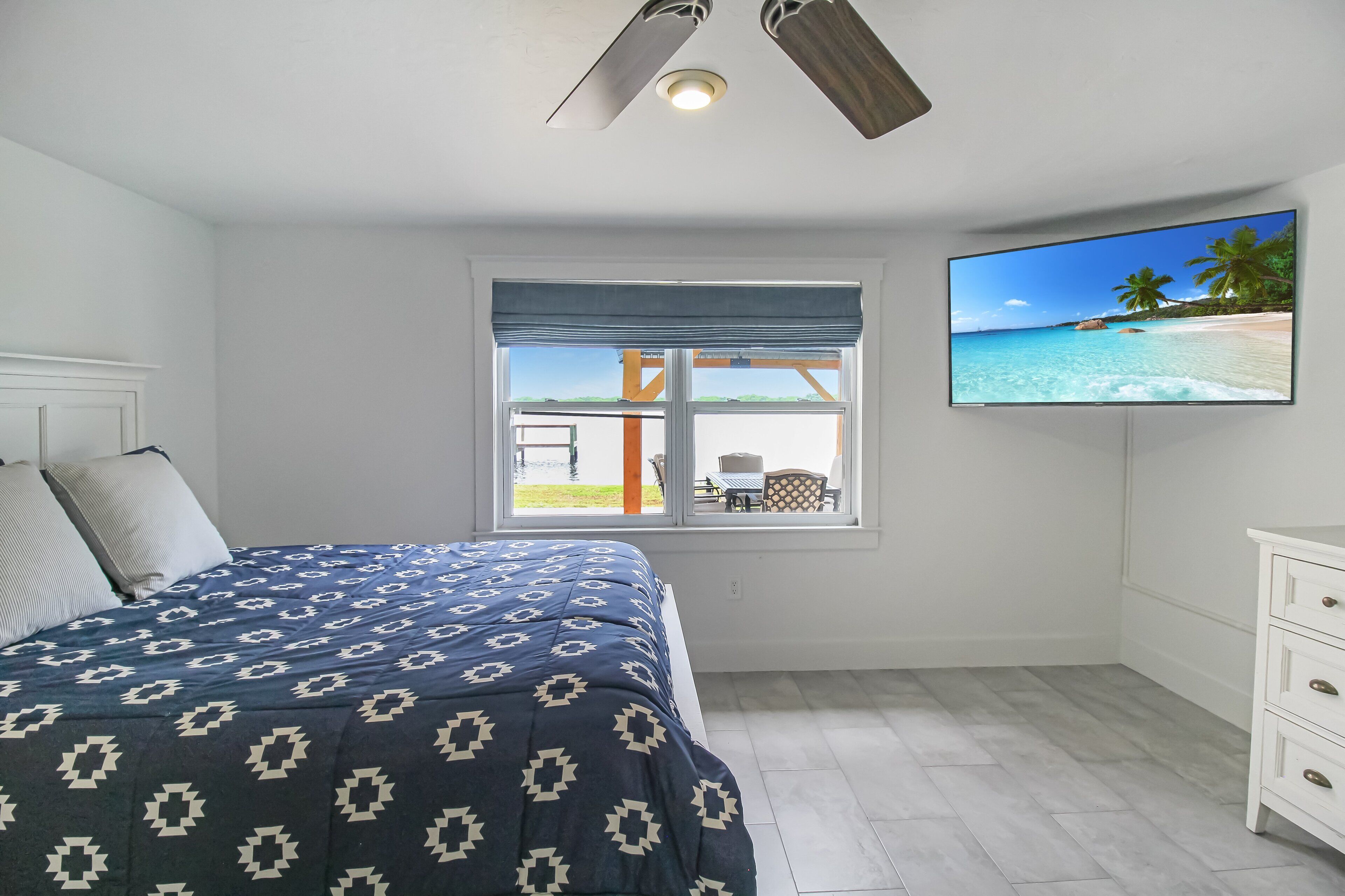 Ein Schlafzimmer mit einem Bett, einem Fernseher, einem Fenster mit Blick auf einen Strand und einem Deckenventilator.