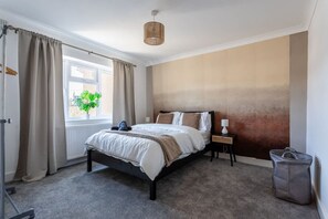 Ein Schlafzimmer mit Bett, Nachttisch, Lampe, einem Fenster mit Vorhängen, einer Pflanze und einer Tasche.