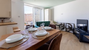 A dining area with a wooden table set for four, a green sofa, and a TV in the background. - Perfect Booking 4 Bed Home With Parking for 2 (Bristol)