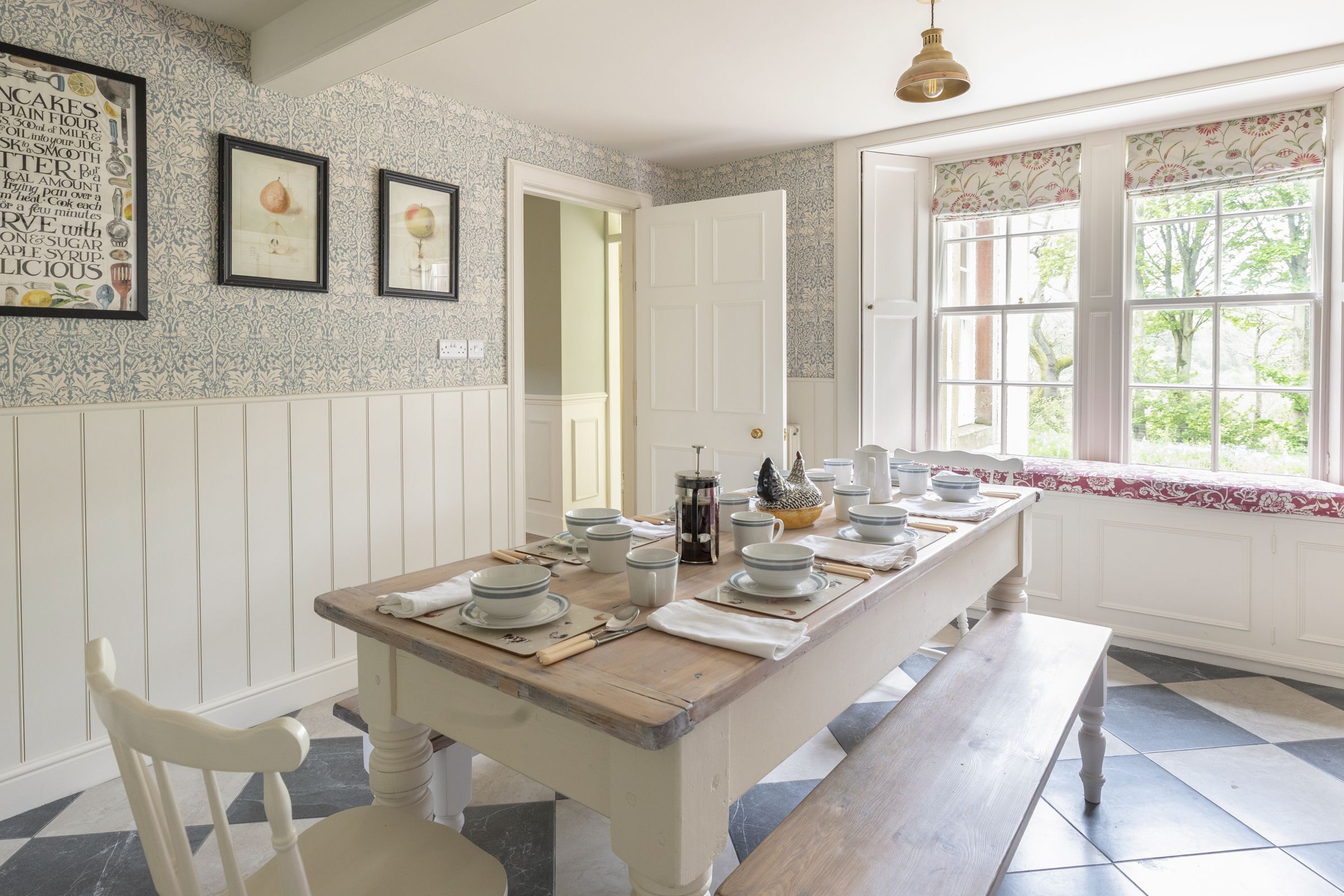 A dining area with a long wooden table set for four, white walls with patterned wallpaper, a window with a view of greenery, and a door leading to another room.