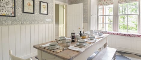 A dining area with a long wooden table set for four, white walls with patterned wallpaper, a window with a view of greenery, and a door leading to another room.