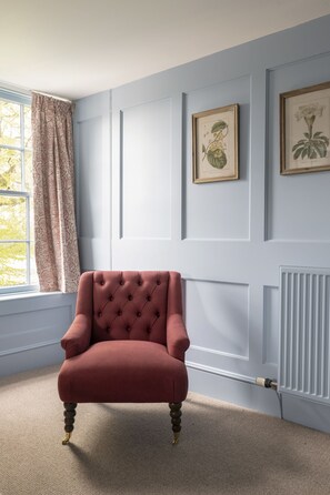 Una habitación con un sillón rojo, una ventana con cortinas y dos grabados botánicos enmarcados en la pared.