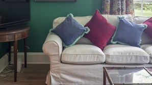 A living room with a green wall, a framed bird illustration, a television, a sofa with colorful cushions, and a patterned curtain. - Sunderland Farmhouse - Sleeping 8 Guests in 4 Bedrooms, Dog Friendly (Galashiels)