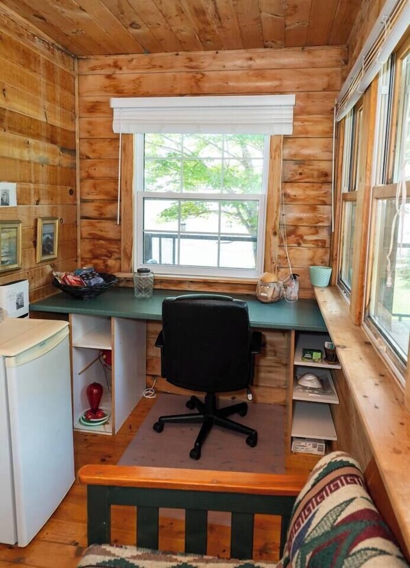 A cabin interior with a wooden desk, a chair, a small refrigerator, and a window with a view of greenery outside.