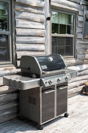 A portable gas grill with four burners, a side burner, and a storage compartment, placed on a wooden deck in front of a log cabin.