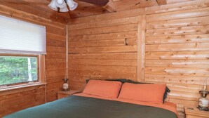 Un dormitorio de cabaña de madera con cama, almohadas, una mesita de noche, una lámpara y una ventana.