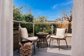 An outdoor deck with wicker chairs and a round table, surrounded by greenery and a clear sky.