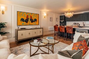 Ein modernes Wohnzimmer mit einer Couch, einem Glastisch, einer Essecke mit Stühlen, einer Küche mit dunklen Schränken und einem großen Bild an der Wand.