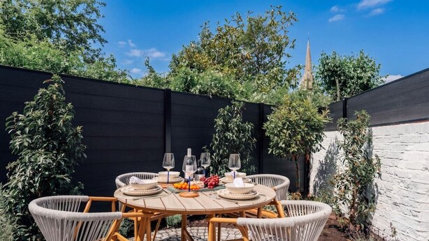 A round wooden table with six wicker chairs, surrounded by a black fence, with a white brick wall and greenery in the background. - Beautiful Fulham Apartment with Aircon (Greater London)