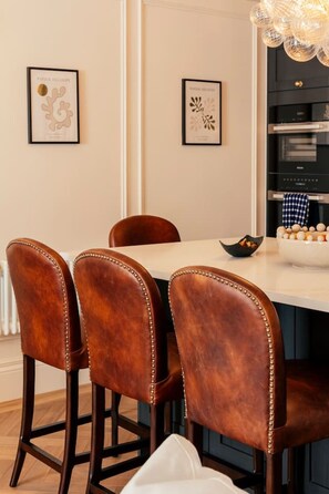 Ein Essbereich mit einem weißen Tisch, vier braunen Polsterstühlen mit Perlenstickkanten und gerahmten Kunstwerken an der Wand.