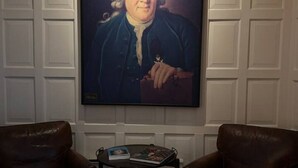 Ett rum med ett stort porträtt på väggen, två läderfåtöljer och ett litet bord med böcker.